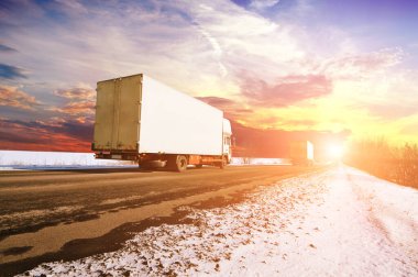 Two white box trucks with space for text on the countryside winter road with snow against sky with sunset clipart