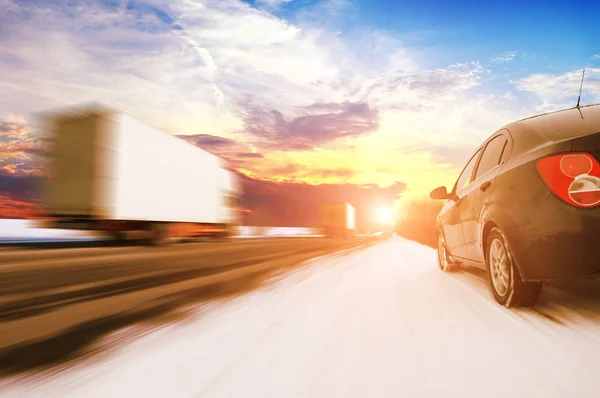 移動中の白いトラック2台と田舎の冬の道路上の黒い家族の車の抽象的な背景明るい日没と空に対する雪と動き — ストック写真