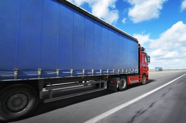 Büyük kırmızı kamyonet ve mavi römork. Yazı için yeri var. Kırsal yolda bulutlu gökyüzüne doğru hızla ilerliyorlar.