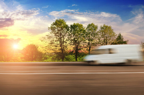 White big van driving fast on the countryside road in motion with green trees and bushes against sky with sunset
