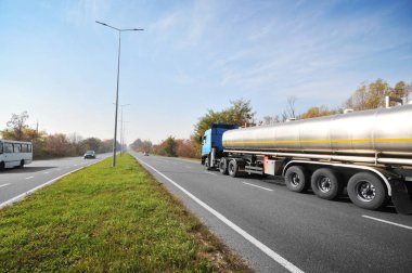 Büyük metal yakıt tankeri diğer arabalarla birlikte mavi gökyüzüne karşı ağaçlı yolda yakıt taşıyor.