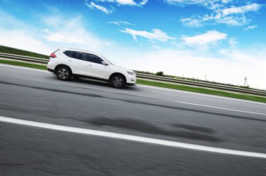 Kırsalda asfalt yolda beyaz bulutlu mavi gökyüzüne doğru hızla giden beyaz bir çapraz araba.