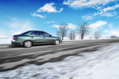 Araba kış kırsalında hızla gidiyor. Kar bulutlu gökyüzüne çarpıyor.