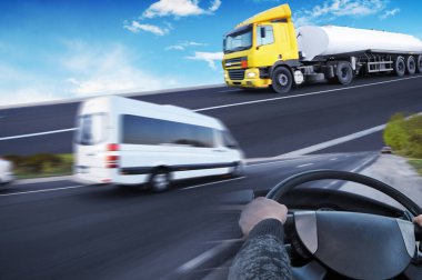 Beyaz minibüs hareket halinde, kamyon direksiyonu ve gökyüzüne karşı büyük tanker kamyonu olan soyut bir arkaplan