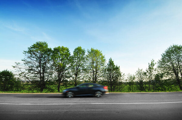 Black sedan car driving fast on the countryside asphalt road with green trees against blue sky