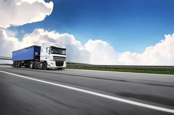 青い空と雲に対して田舎道上の動きで青いトレーラーを持つ大きな白いトラック — ストック写真