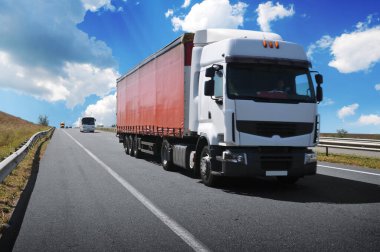 Büyük beyaz kamyon ve kırmızı römork kırsal yolda diğer araçlarla birlikte bulutlu mavi gökyüzüne karşı