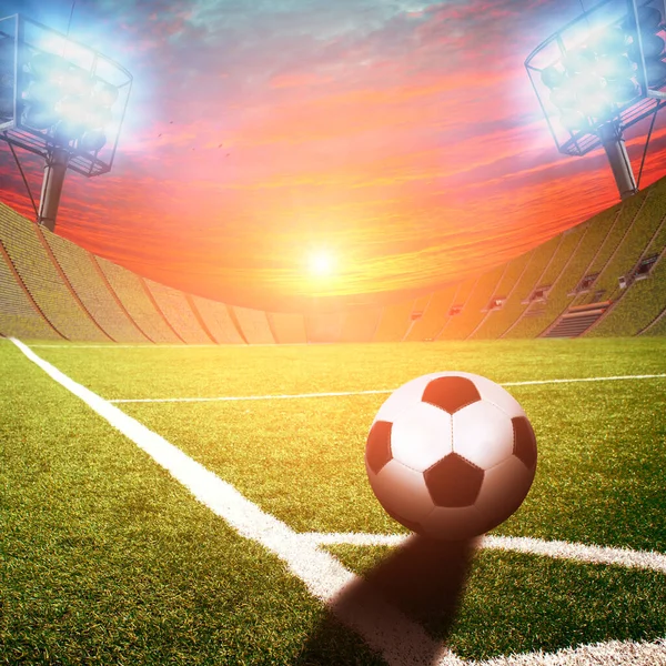 Football stadium with black and white ball on corner of field with green grass and bright lights with flashes against sky with sunset