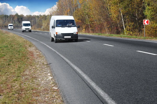 Three white commercial vans on countryside road with forest against blue sky with clouds