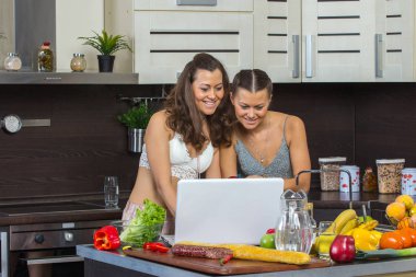 Dizüstü bilgisayar, mutfakta ayakta bakarak iki çekici kadın. 