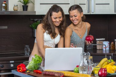 Dizüstü bilgisayar, mutfakta ayakta bakarak iki çekici kadın. 