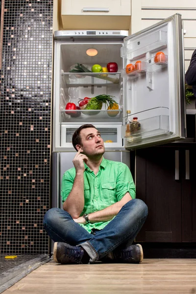 Молодой человек сидит перед холодильником — стоковое фото