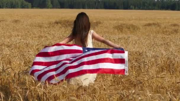 Belleza chica corriendo en el campo de trigo amarillo con bandera nacional de EE.UU. . — Vídeo de stock