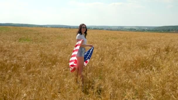 Menina de beleza correndo no campo de trigo amarelo com bandeira nacional dos EUA. Mulher feliz ao ar livre. Colheita — Vídeo de Stock