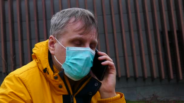 Sick man in medical mask speaks on the phone while sitting on a park bench — Stock video