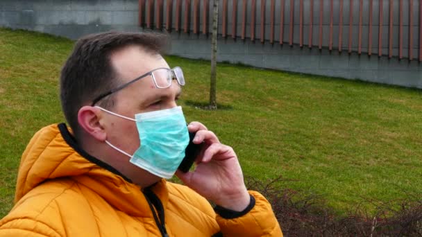 Больной человек в медицинской маске говорит по телефону, сидя на скамейке в парке — стоковое видео