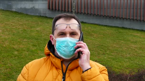 Sick man in medical mask speaks on the phone while sitting on a park bench — Αρχείο Βίντεο
