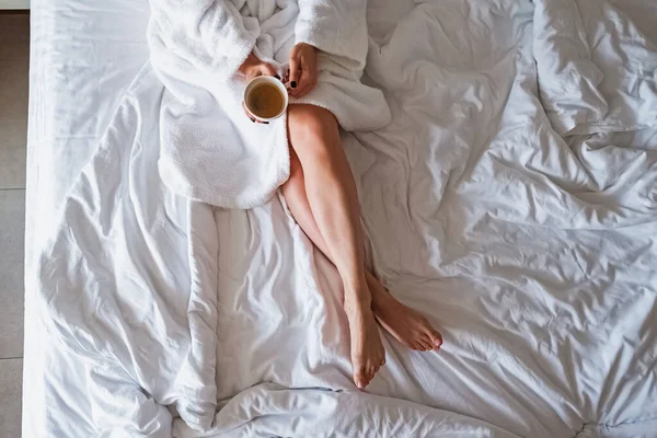 Невизначена жінка в білому халаті, насолоджуючись ранковою кавою, сидячи на своєму незробленому ліжку . — стокове фото