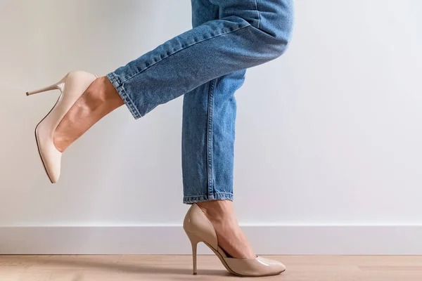 Woman dressed in denim pants and high heels standing near the white wall with one leg raised.