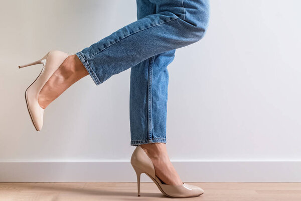 Женщина в джинсовых брюках и на высоких каблуках стояла возле белой стены с одной поднятой ногой
.