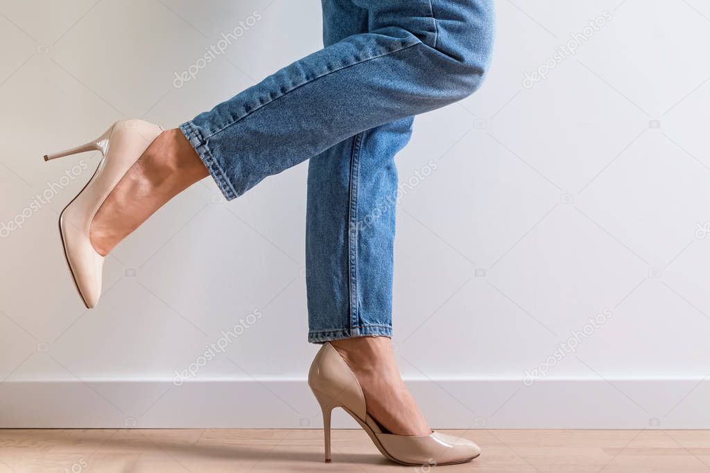 Woman dressed in denim pants and high heels standing near the white wall with one leg raised.