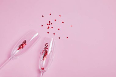 Pembe arka planda kırmızı kalp şeklinde konfeti damlatan iki şampanya kadehi..