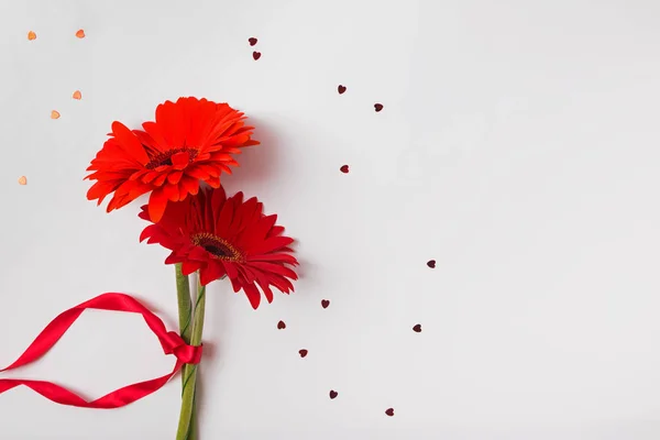 Két piros gerbera virág piros szalaggal és szív alakú csillám konfetti fehér háttér másolási hely. — Stock Fotó