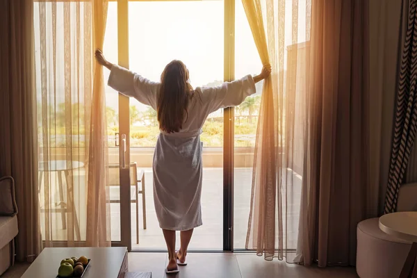 Young woman wearing white bathrobe opening curtains in luxury hotel room — Stock Photo, Image