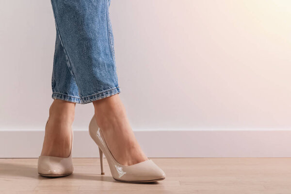 Женские ноги в джинсах и туфлях на высоком каблуке стоят на полу возле белой стены
