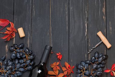 Kırmızı üzümler, bir şişe şarap ve siyah ahşap arka planda yapraklar..