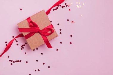Pembe arka planda kırmızı pruvayla dekore edilmiş hediye kutusu. Kalp şeklinde simli konfeti..