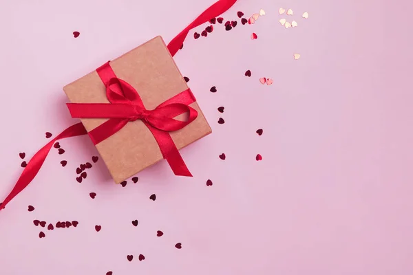 Ajándék doboz díszített piros íj rózsaszín háttér szív alakú csillogó konfetti. — Stock Fotó