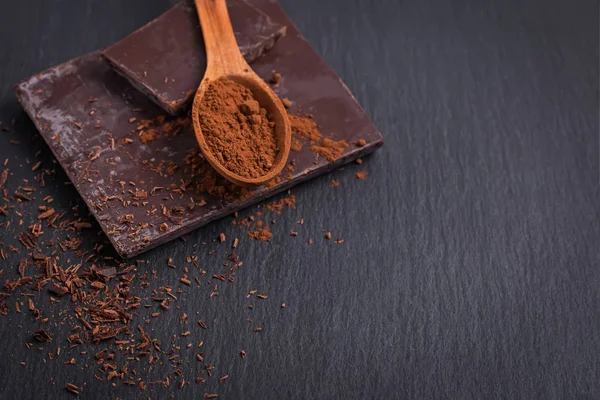 Bar of natural chocolate and cocoa powder in a wooden spoon on black stone bacgkround — Stock Photo, Image