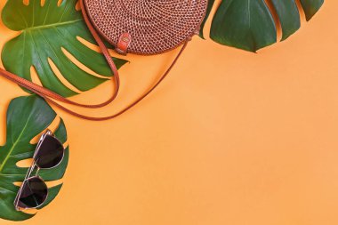 Yuvarlak şekilli rattan çanta ve güneş gözlüğü sarı arka plandaki canavar yapraklarının üzerinde duruyor.