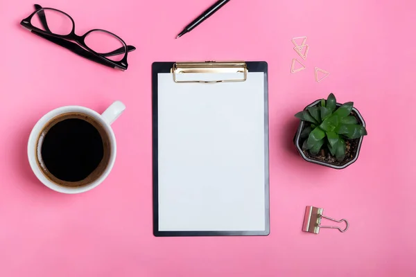 Кліпборд з чистим папером на рожевому столі з кавою, окулярами та соковитими рослинами — стокове фото