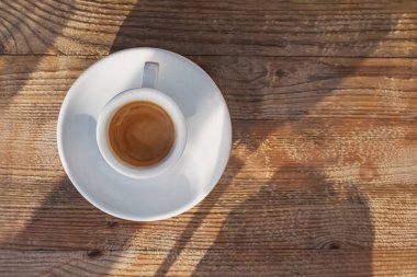 Bir fincan kahve, güneş ışığında ahşap masanın üzerinde duruyor. Gölgeler düşüyor.