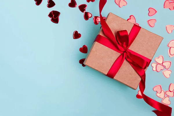Подарункова коробка з червоною стрічкою та конфітті у формі серця на синьому фоні . — стокове фото