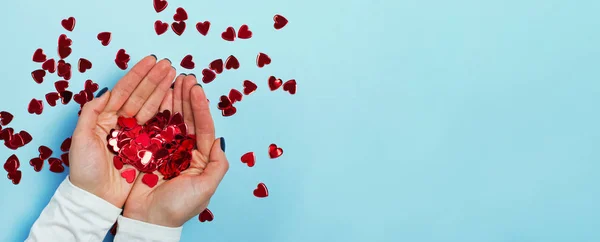 Mains féminines tenant des confettis rouges étincelants en forme de cœur sur fond bleu — Photo