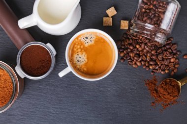 Bir fincan kahve ve espresso makinesinin filtre tutacağı ve siyah masada kızarmış kahve çekirdekleri.