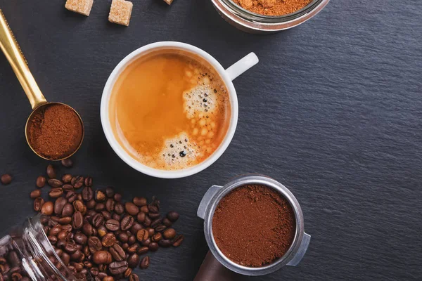 Uma xícara de café e filtro titular da máquina de café expresso e com grãos de café torrados espalhados na mesa preta — Fotografia de Stock