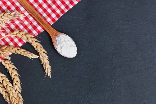 Wooden spoon with flour, wheat ears and red checkered cloth on black table — Stock Photo, Image