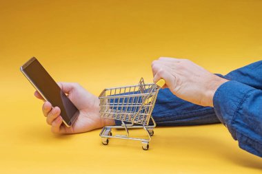 Sarı arka planda küçük alışveriş arabası ve akıllı telefon tutan eller..