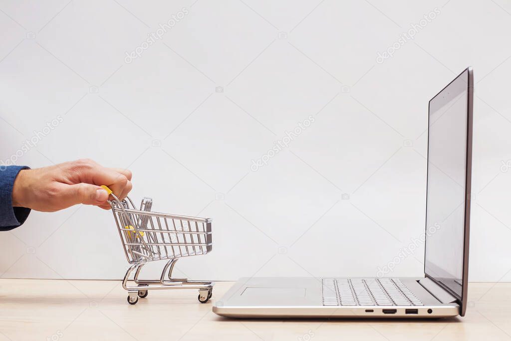 Hand holding mini shopping trolley near the opened laptop on yellow background.