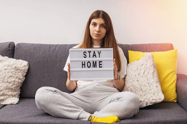 Woman sitting on the sofa in her living room holding lightbox with message Stay Home