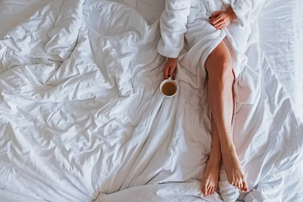 Onherkenbare vrouw in witte badjas zittend op het bed met een kop koffie in haar hand — Stockfoto