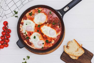 Shakshuka yumurta kabı, beyaz ahşap masada siyah dökme demir bir tavada domates sosu.