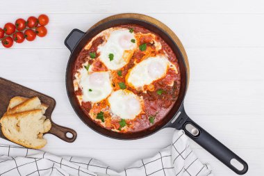 Shakshuka yumurta kabı, beyaz ahşap masada siyah dökme demir bir tavada domates sosu.
