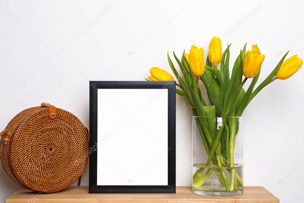 Empty frame standing on small wooden table with yellow tulips and straw bag