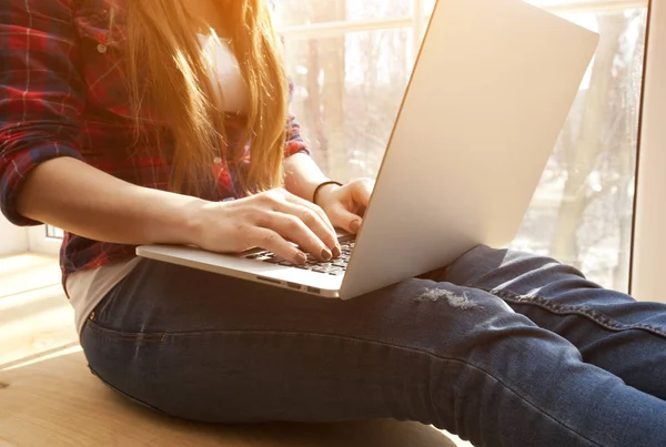 Die junge Frau sitzt auf der Fensterbank und arbeitet am Laptop — Stockfoto