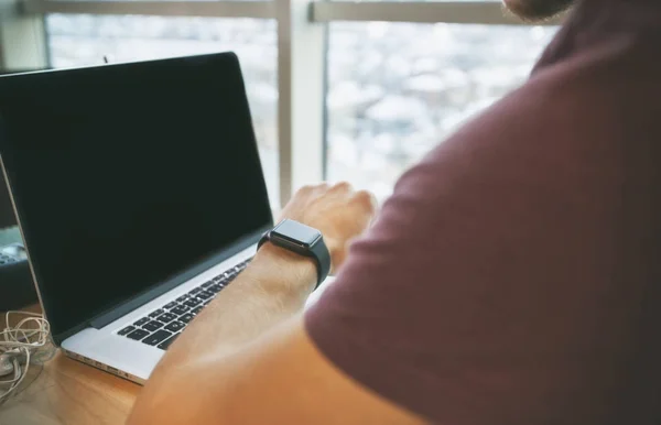 O jovem sentado no laptop e olhando para o smartwatc — Fotografia de Stock